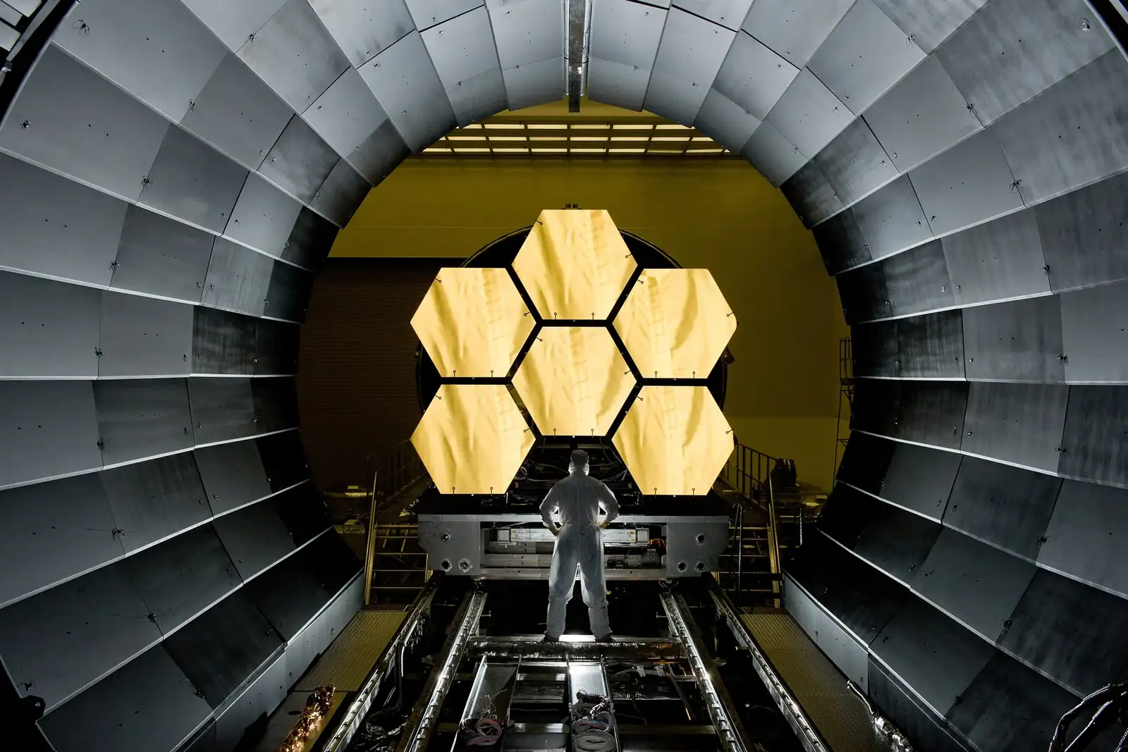 Un ingénieur, de dos, observe six segments de miroir hexagonaux dorés montés sur un banc d'essai dans une grande chambre d'essais