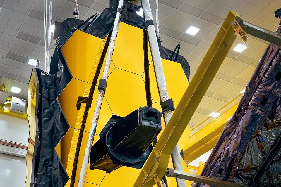 Des techniciens en combinaison blanche autour d'un grand miroir doré de télescope dans une salle d'intégration